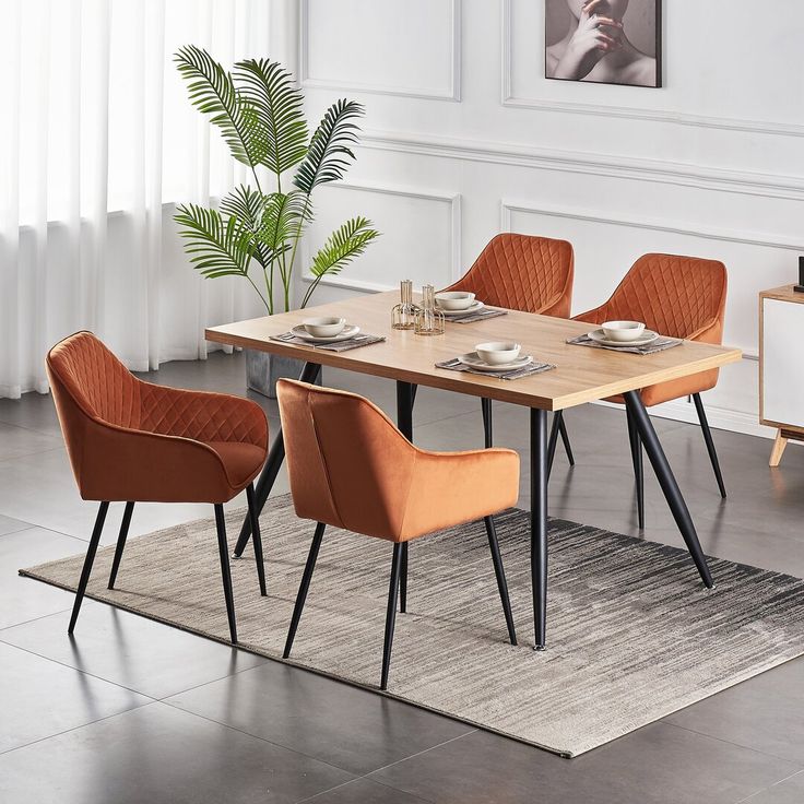 Modern wooden dining table with black metal legs, paired with elegant orange velvet chairs in a stylish dining room with white panel walls, a large window, and indoor plants.