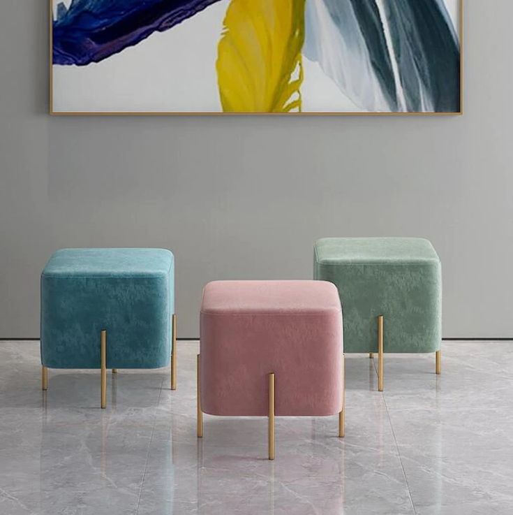 Three pastel-colored square velvet ottomans with gold legs set against a minimalist gray wall with modern artwork.