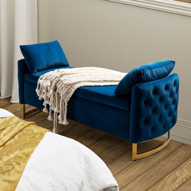 Elegant blue velvet bench with tufted sides, gold rocking legs, and plush pillows, placed near a bed with a cozy knit throw.