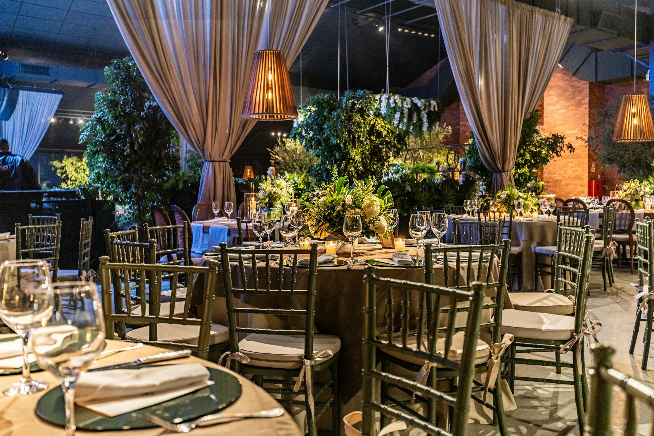A beautifully decorated banquet hall with elegant table settings, lush greenery, and warm ambient lighting. The dining area features classic wooden chairs with cushions, round tables with fine dining setups, and stylish hanging lamps, creating a luxurious and inviting atmosphere.