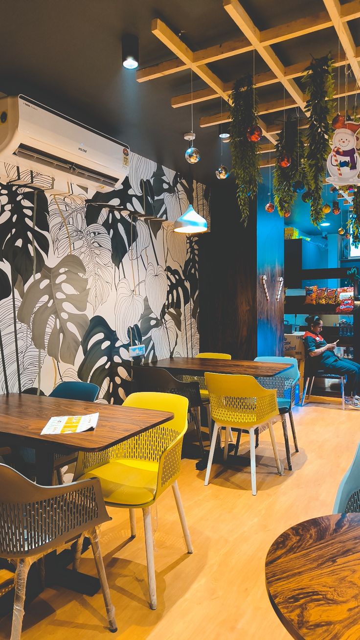 Colorful café with yellow and grey woven plastic chairs, wooden tables, tropical leaf wall art, and festive ceiling decor