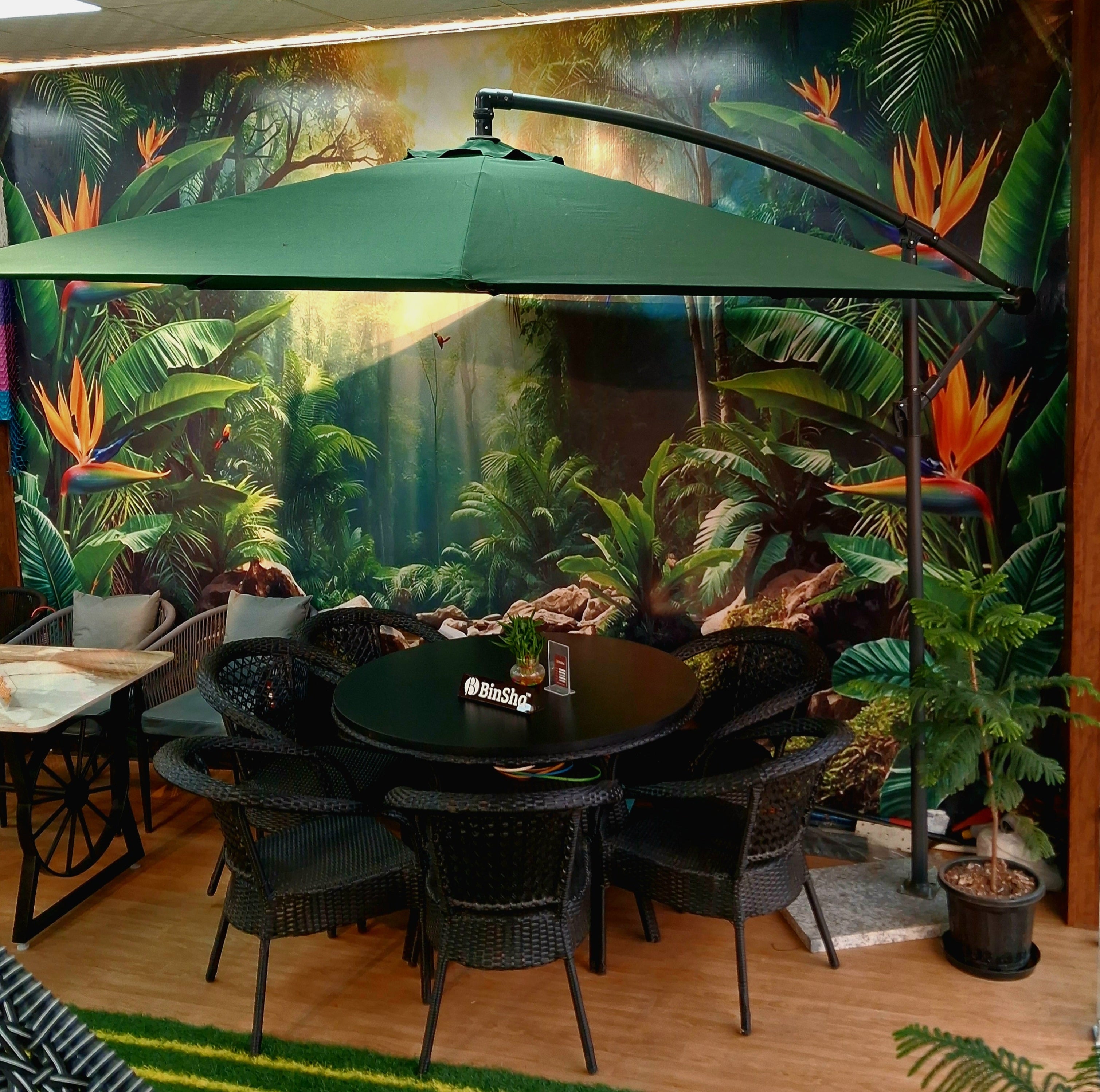Dining area with a large green umbrella, table, and chairs against a tropical mural.