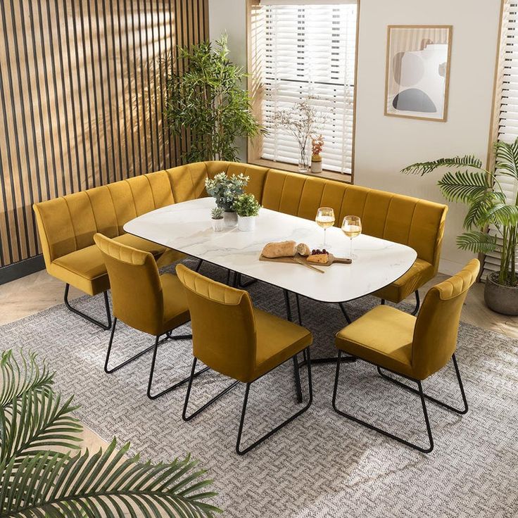 Modern dining set with a white marble table, mustard velvet chairs, and an L-shaped bench in a stylish dining room with wooden panel walls and indoor plants.