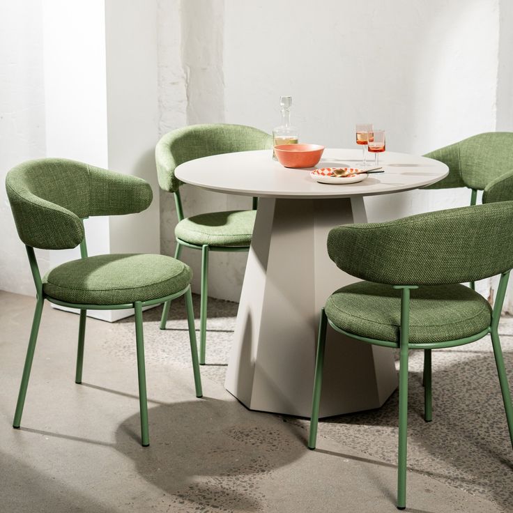 Modern dining set with round white table and four green upholstered chairs in a minimal interior.