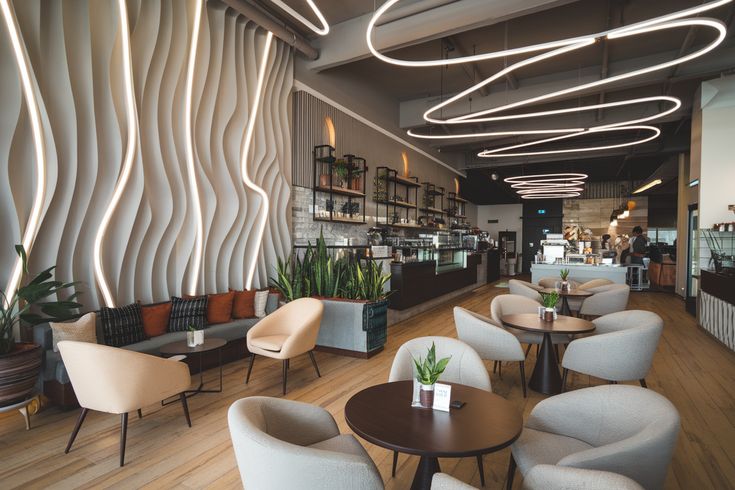 Modern café interior with beige lounge chairs, dark round tables, and a wavy ambient-lit wall panel with hanging LED ceiling lights