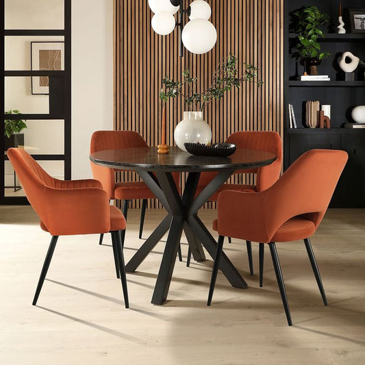 Modern round black dining table with an X-shaped base, surrounded by four orange velvet chairs in a stylish dining room with wood panel walls and contemporary décor.