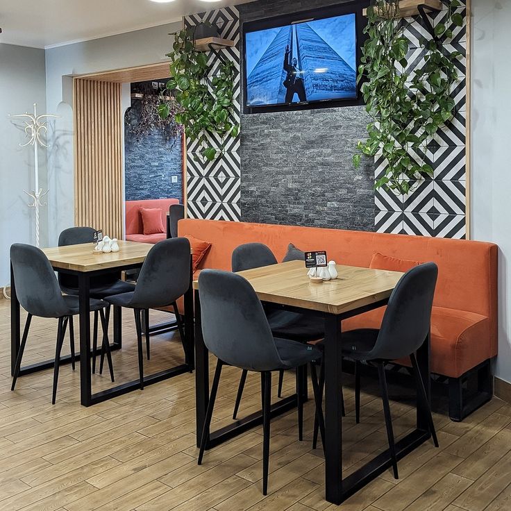 Modern café with orange bench seating, black cushioned chairs, wooden tables, and a black-white geometric wall design accented with green hanging plants.

