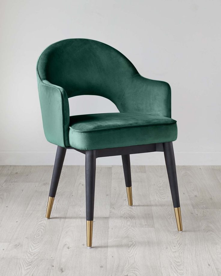 Emerald green velvet chair with curved open back, black tapered legs, and gold tip accents in a minimalist interior.