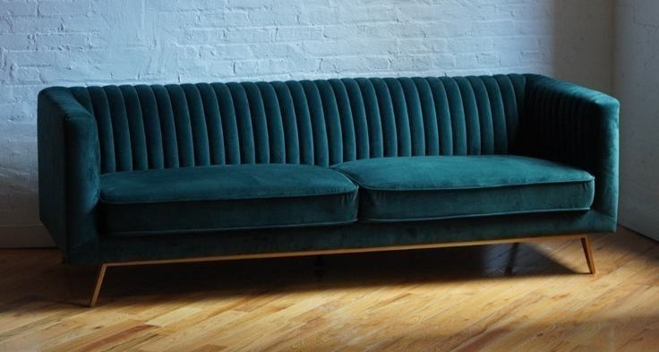 Teal velvet Chesterfield sofa with gold legs against a white brick wall in a minimalist interior.