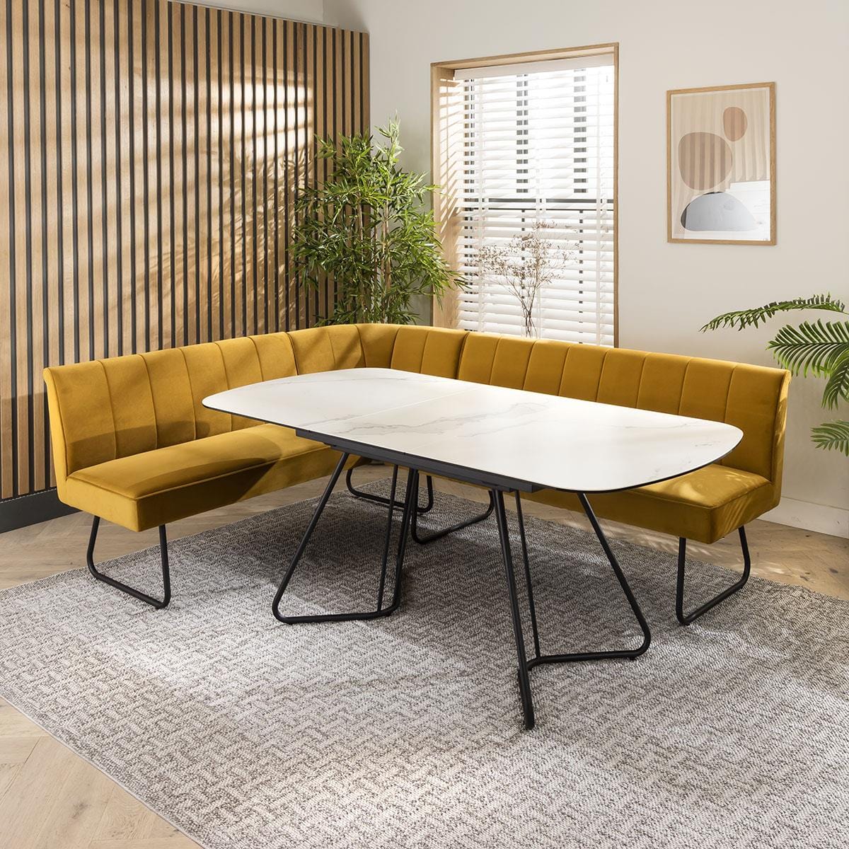 Modern dining set with a white marble table, mustard velvet chairs, and an L-shaped bench in a stylish dining room with wooden panel walls and indoor plants.